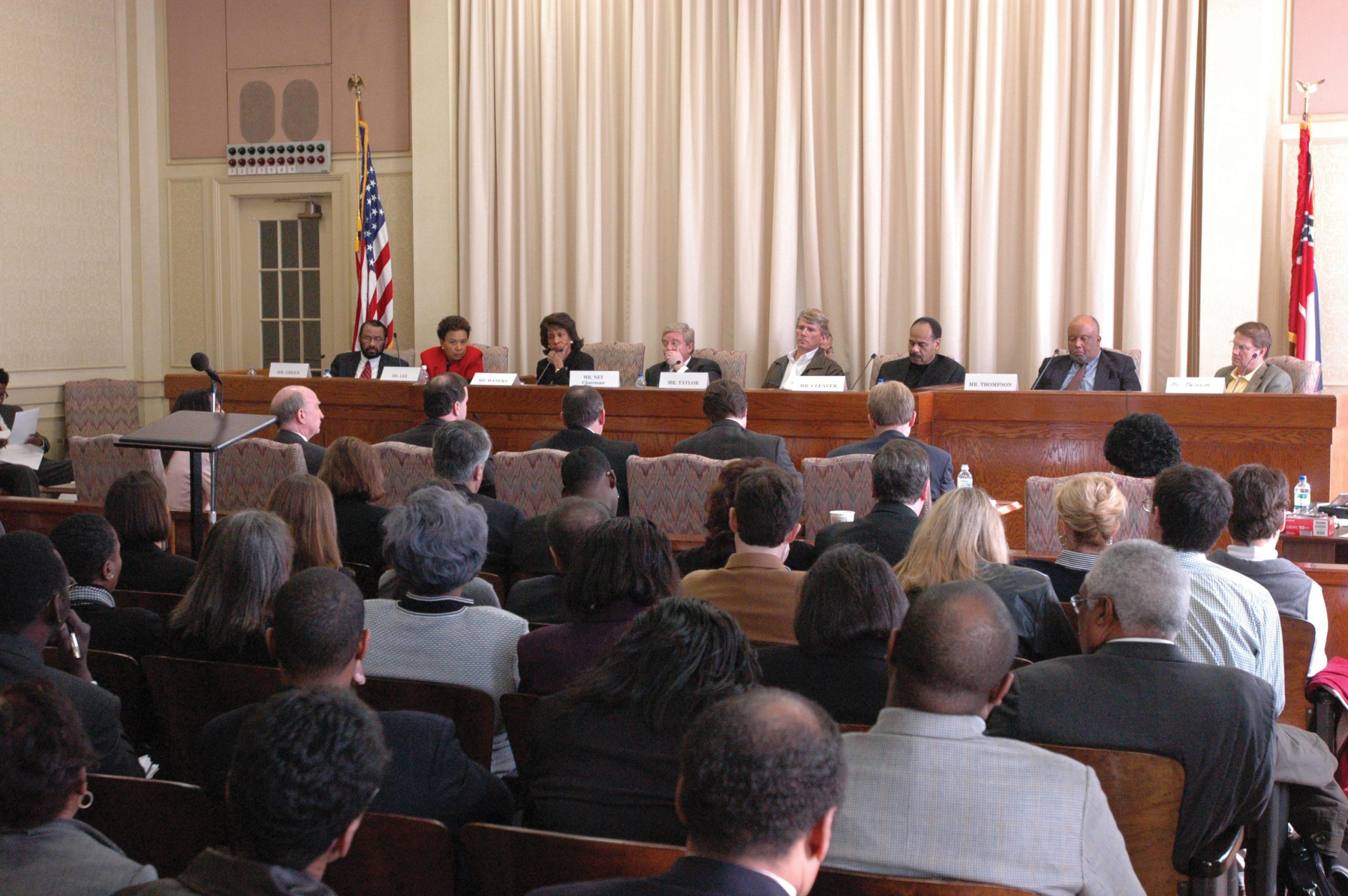 "A large room with a panel of nine people seated at a long table in front of beige curtains, facing an audience. Behind the panelists are the American flag on the left and another flag on the right. The audience is seated in rows of chairs, and a podium with a microphone is positioned to the left side of the image."