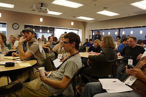 "A group of people are seated in a conference room, attentively listening to a speaker. The room has several round tables with chairs around them. Some attendees are holding cups and notepads, while others have name tags hanging from their necks. The ceiling has recessed lighting and there is a clock on the wall. A projector is mounted on the ceiling, indicating that presentations may be part of the event." This image captures a conference or seminar setting where participants are engaged in listening to a speaker or presentation, highlighting an educational or professional environment where knowledge sharing and networking might be taking place.