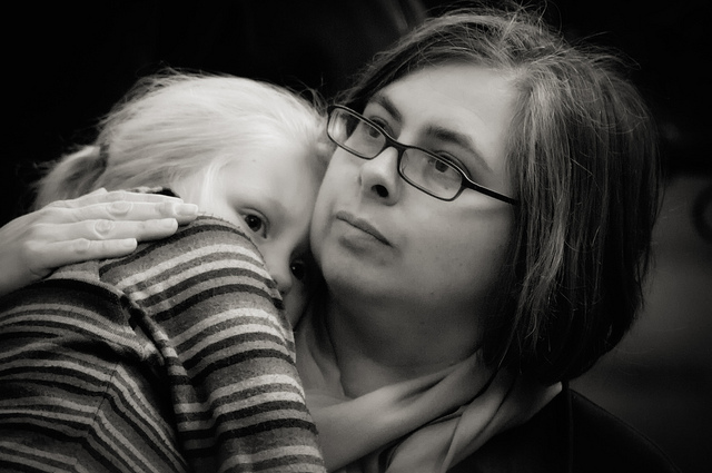 "A black and white photograph shows a young child with light hair resting their head on the shoulder of an adult. The child's face is visible, looking slightly to the side. The child is wearing a striped shirt and appears to be comforted by the adult's embrace."