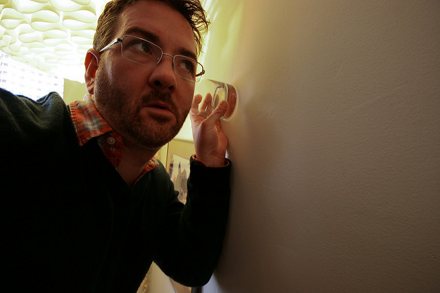 "A person is holding a glass against a wall and leaning their ear to the glass, as if trying to listen through the wall. The person is wearing glasses, a dark sweater, and a checkered shirt. The background shows part of an interior space with patterned ceiling tiles and some framed pictures on the wall."