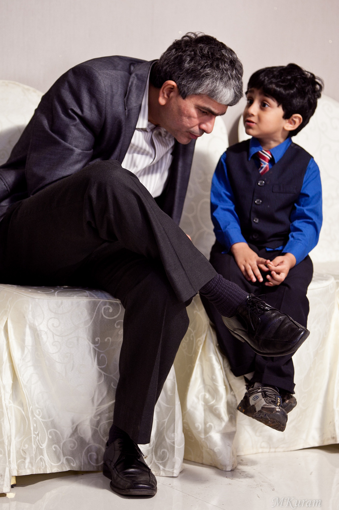 "An adult and a child are sitting on a white couch. The adult, dressed in a dark suit, is leaning forward with legs crossed and appears to be talking to the child. The child's face is blurred for privacy, and the child is wearing a blue shirt with a dark vest and tie. Both individuals are seated closely together, suggesting an intimate or important conversation."
