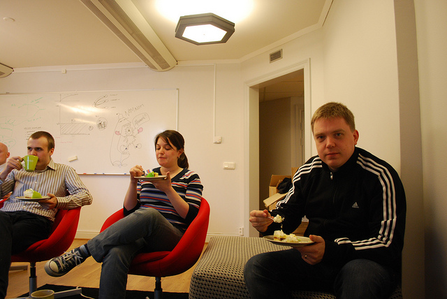 "Three people are seated on red chairs in a casual room, eating food. A whiteboard with various drawings and notes is visible in the background."