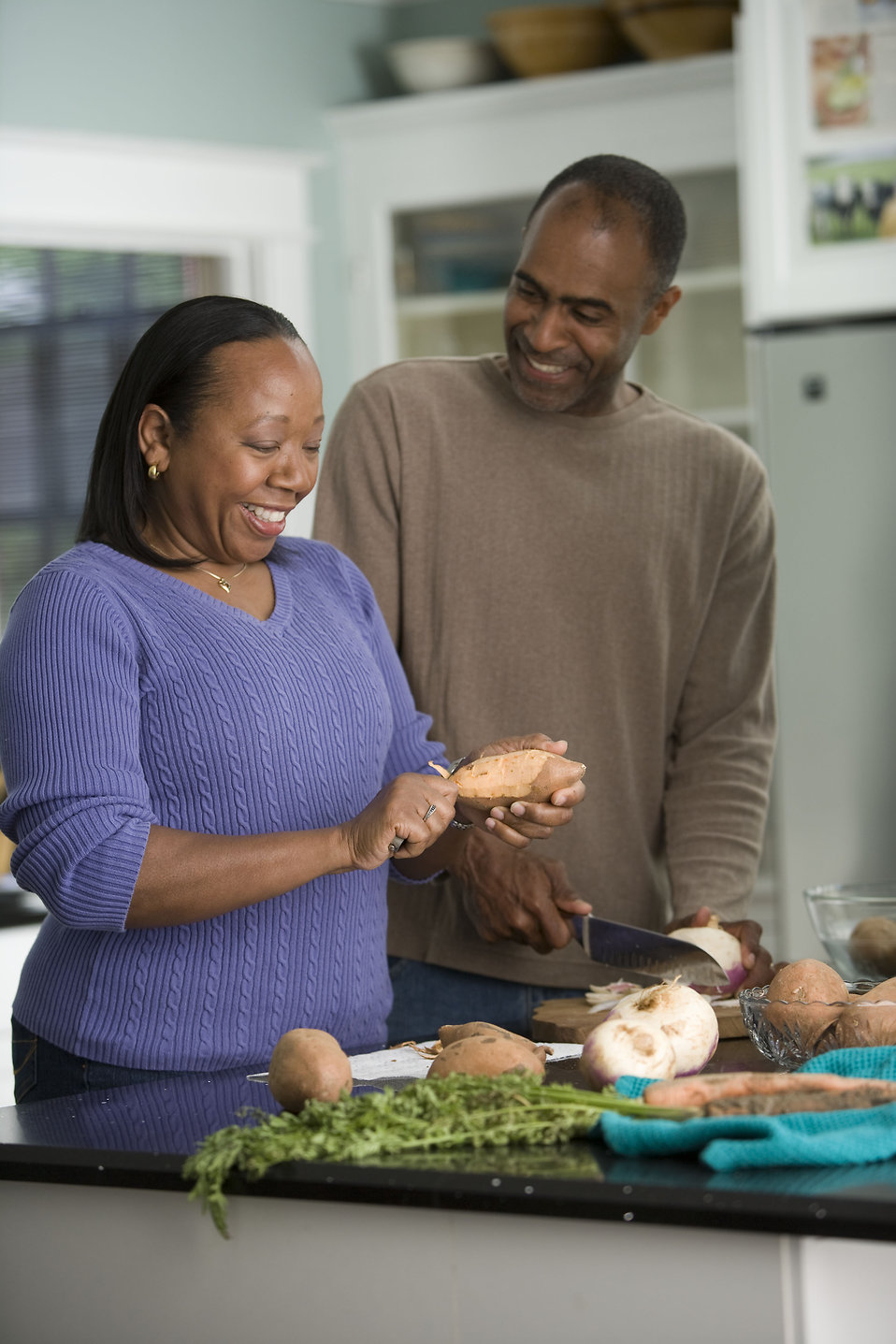 "Two people are standing in a kitchen preparing food. The person on the left, wearing a purple sweater, is peeling a sweet potato, while the person on the right, in a brown sweater, is cutting another sweet potato with a knife. Various vegetables, including sweet potatoes and turnips, are spread out on the counter. The kitchen features light blue walls and white cabinets with glass doors."