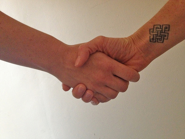 "Two hands are shaking. The hand on the right has a tattoo of an intricate geometric pattern on the wrist. The background is plain and light-colored."