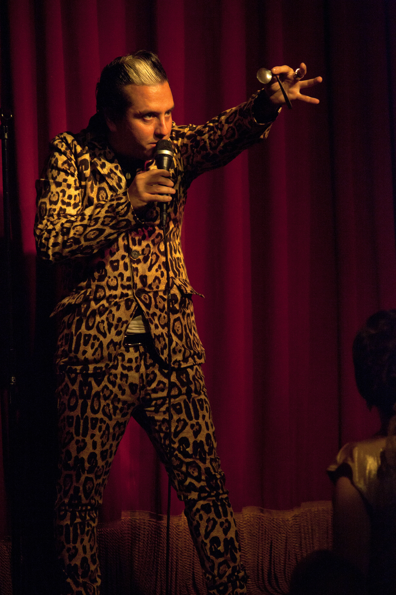 "A person in a leopard print suit stands on stage in front of a red curtain, holding a microphone in one hand and gesturing with the other. Another person is partially visible sitting in the foreground on the right side."