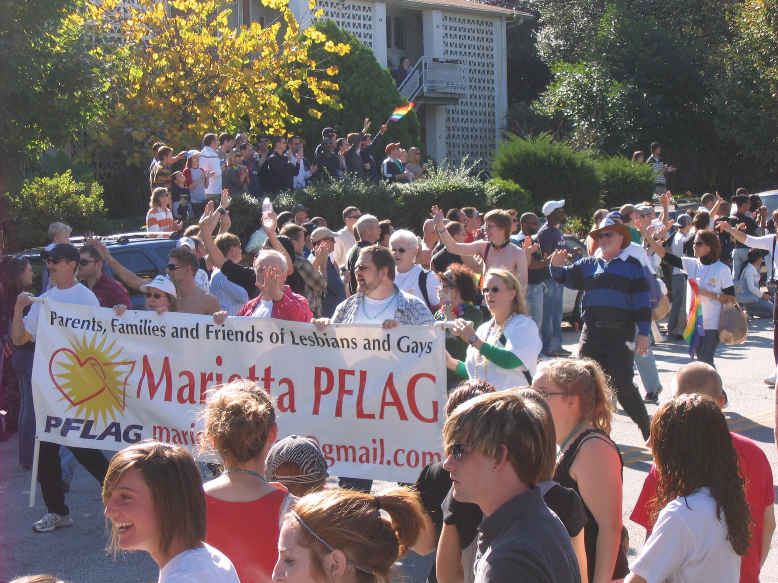 "A large group of people participating in a parade or rally, holding a banner that reads 'Parents, Families and Friends of Lesbians and Gays Marietta PFLAG.' The diverse crowd includes individuals of various ages, some carrying rainbow flags. The event is outdoors on a sunny day, with trees and buildings in the background."