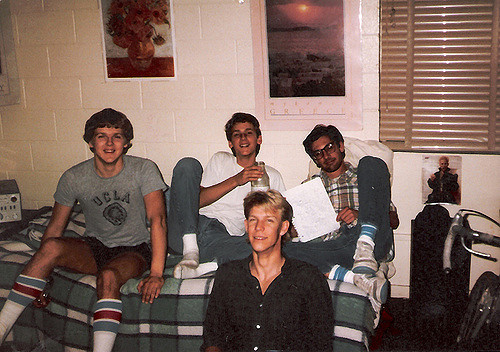 "Four people are sitting on a bed in a room with white brick walls. The person on the left is wearing a gray UCLA t-shirt and striped socks. The second person is holding a bottle, the third is holding a piece of paper, and the fourth, wearing a black shirt, is seated in front of them. Posters are on the wall behind them, and blinds partially cover a window."