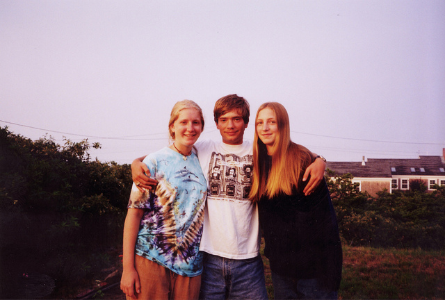 "Three individuals standing outdoors with their arms around each other. The person on the left is wearing a tie-dye shirt and beige pants, the person in the middle is in a white graphic t-shirt and blue jeans, and the person on the right is wearing a black long-sleeve shirt and blue jeans. Bushes and houses are visible in the background."