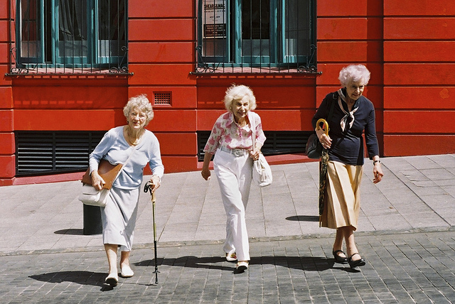 "Three elderly individuals walking on a cobblestone street in front of a bright red building with blue window frames. The person on the left uses a cane and carries a bag. The person in the middle, wearing white pants, holds onto the arm of the person on the right, who is carrying a patterned bag."