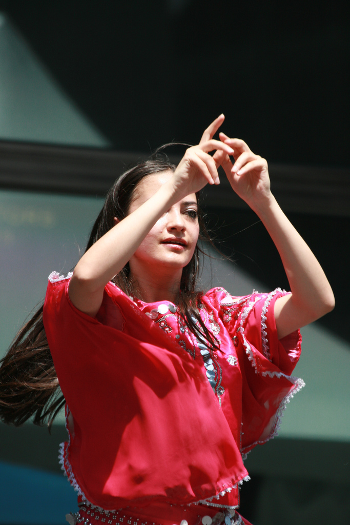 "A person wearing a red garment with white decorative trim is using American Sign Language. The background appears to be an outdoor setting with some visible structures."
