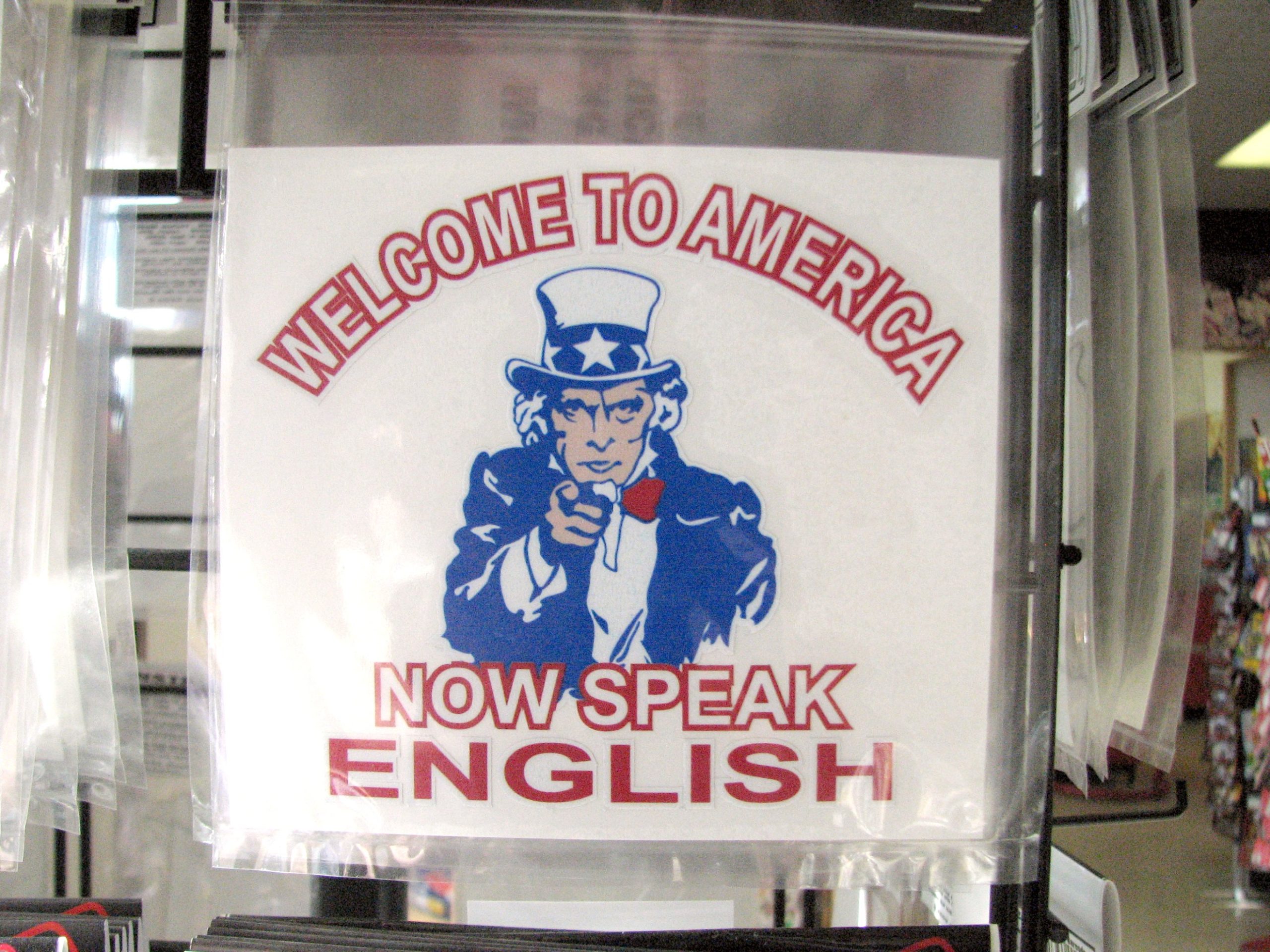 "A sign with an image of Uncle Sam pointing forward. The text above the image reads 'WELCOME TO AMERICA' in red capital letters, and below the image, it reads 'NOW SPEAK ENGLISH' also in red capital letters."