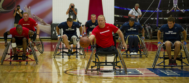 "A group of athletes in wheelchairs are playing basketball on an indoor court. The players are wearing uniforms with 'NAVY' and 'COAST GUARD' written on them, and one player in a red shirt is holding a basketball. In the background, there are photographers capturing the event."