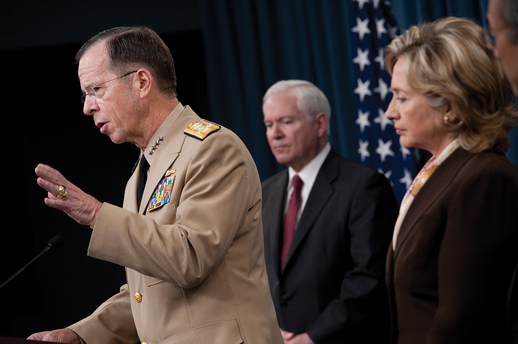 "Four people stand in front of a dark blue curtain with an American flag in the background. The person on the left, wearing a beige military uniform with medals and insignia, speaks into a microphone. The other three individuals are in formal attire, including suits and a blazer."