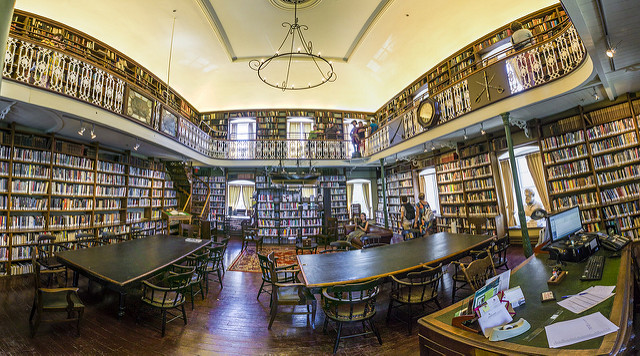 "A spacious, two-story library with tall bookshelves filled with books. The library features a high ceiling with a chandelier, ornate railings on the upper level, and several tables and chairs on the ground floor. A computer workstation is visible in the foreground, and people are browsing books on both levels."