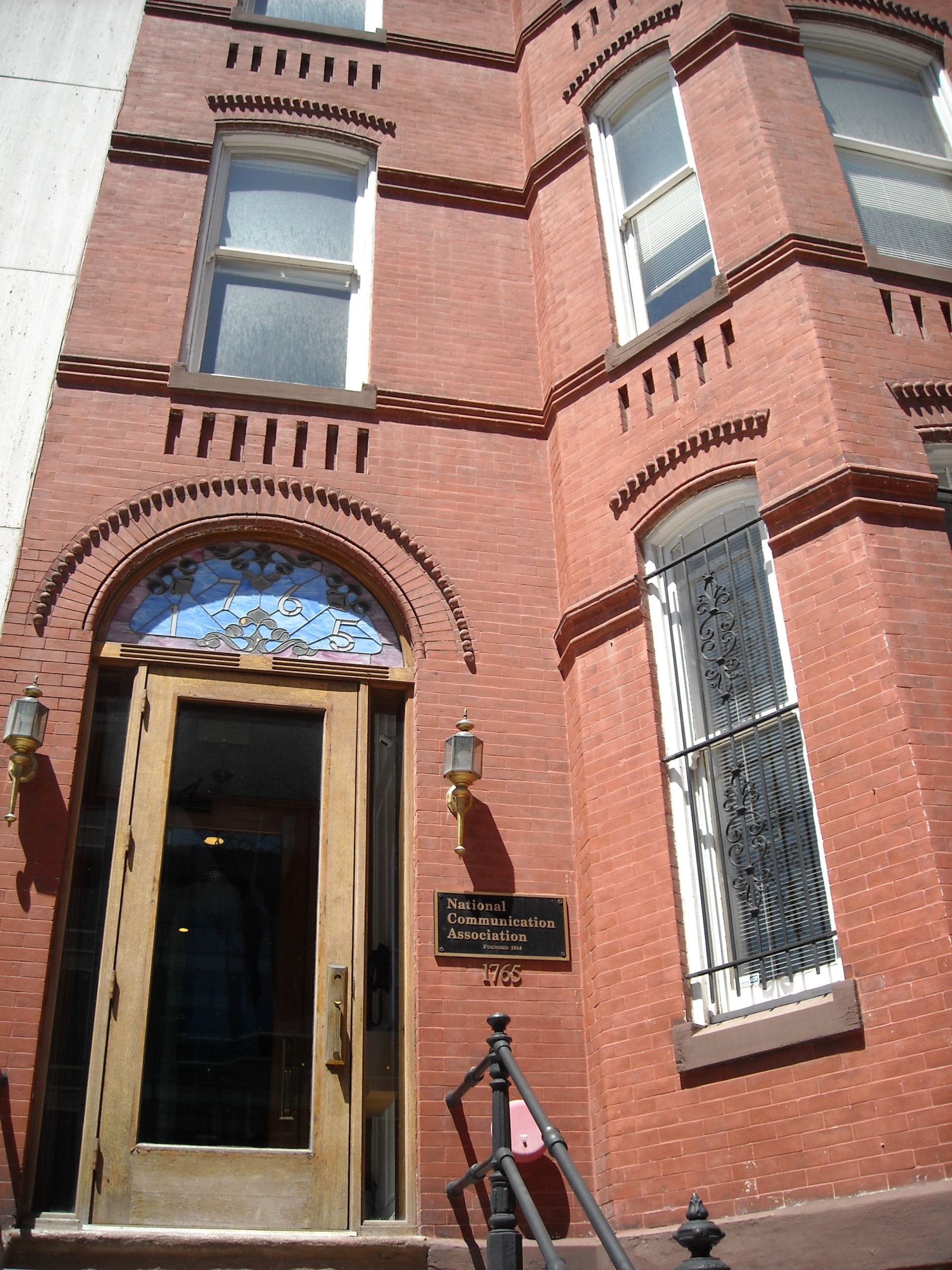 "Entrance to a brick building with an arched doorway and a decorative stained glass window above a wooden door with glass panels. Two lantern-style lights flank the door. A black sign with gold text to the right reads 'National Communication Association' with the number '1765' below. The building features tall windows with white frames and decorative brickwork above them."