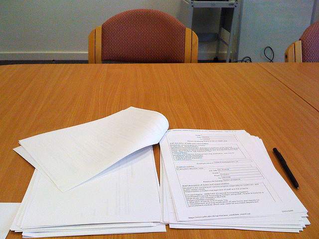 "A wooden table with a stack of papers on the right, including printed text and forms, and a black pen beside them. More papers are scattered on the left, some with curled edges. In the background, two red-patterned upholstered chairs and a metal cart with electronic equipment are positioned against a white wall."