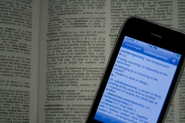 "An open dictionary with an iPhone resting on top. The phone screen displays the definition of the word 'contemporary' from an online dictionary app, including meanings like 'living or occurring at the same time' and 'belonging to or occurring in the present.' The dictionary page beneath shows blurred text with various words and definitions."