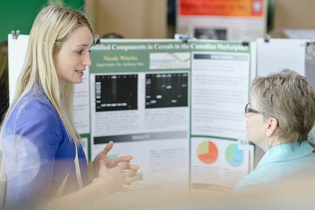 "Two individuals standing in front of a scientific poster titled 'Modified Components in Cereals in the Canadian Marketplace' by Nicole Warkentin from the Department of Food and Human Nutritional Sciences. The poster includes multiple sections with text, a bar graph, and pie charts in various colors representing different data points."