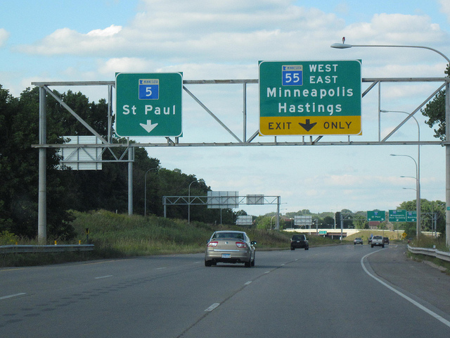 "A highway scene with two overhead road signs. The left sign shows Interstate 5 toward St. Paul with a downward arrow. The right sign shows Interstate 55 with directions to West and East Minneapolis and Hastings, marked 'EXIT ONLY' in yellow. Several cars are on the road, with trees lining both sides and a partly cloudy sky overhead."