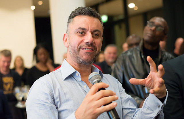 "A person in a light blue button-up shirt is holding a microphone and speaking, with one hand gesturing. The background includes several other people, some blurred, suggesting an indoor event or gathering."