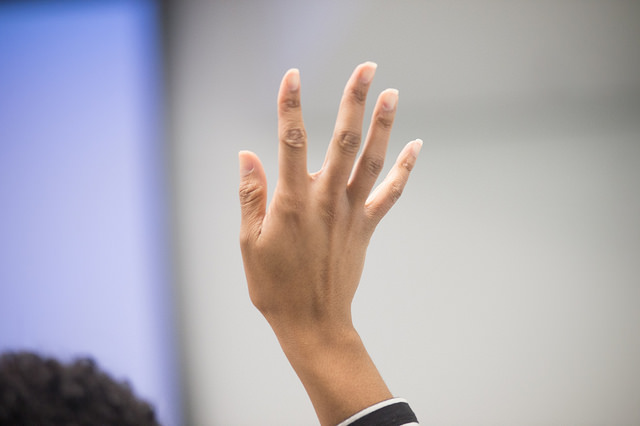 "A close-up of a person's hand raised with fingers spread apart. The background is blurred, showing indistinct colors and shapes."