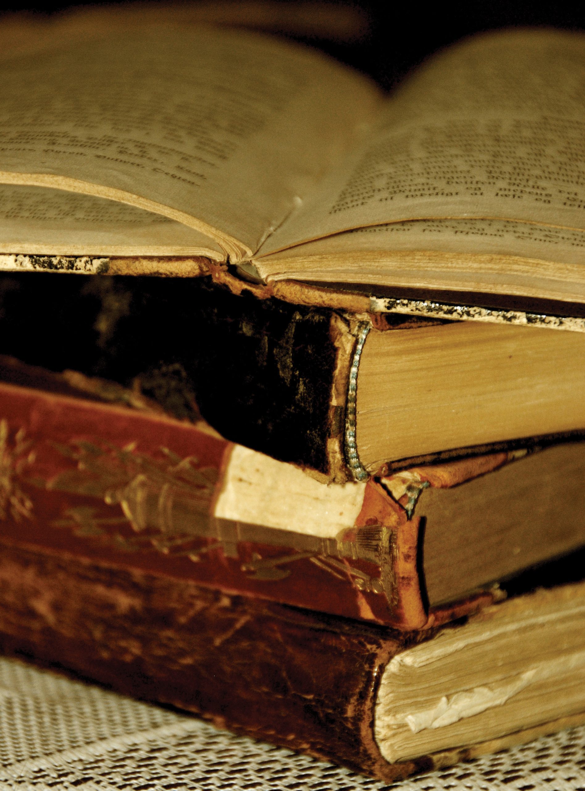 "A close-up of three old, worn books stacked on a lace tablecloth. The top book is open, showing yellowed pages with text. The middle and bottom books are closed, with visible wear on their spines and edges."