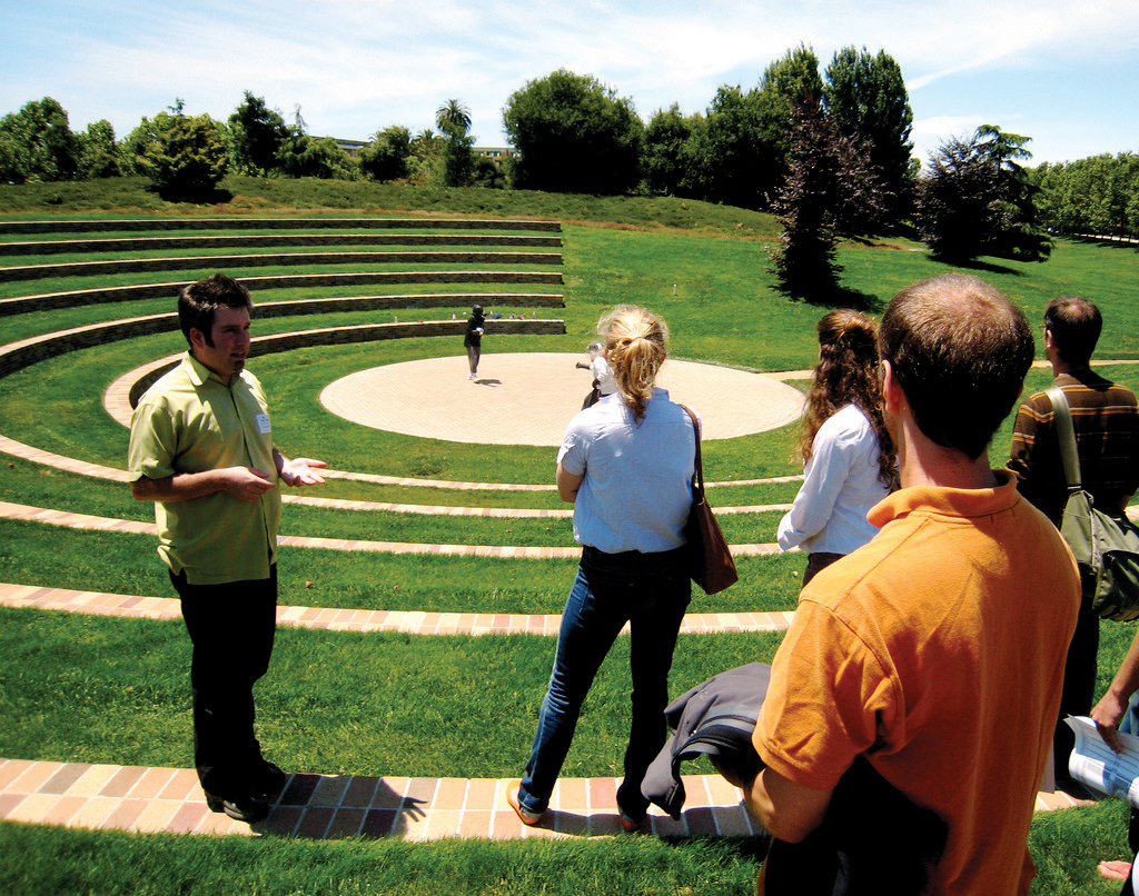 "A group of people stand on the steps of an outdoor amphitheater with a circular stage at the center. One person is on the stage, while others are scattered around the steps—some facing the stage, others interacting with each other. The amphitheater is surrounded by green grass and trees under a mostly clear sky with a few clouds."