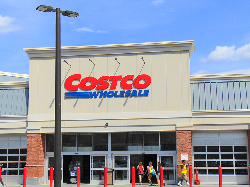 The exterior entrance of a Costco Wholesale store on a sunny day. The large red and blue Costco logo is prominently displayed above the entrance. A few shoppers, including one wearing a safety vest, are walking toward the sliding glass doors. Red safety bollards line the front of the store, and a tall black streetlight stands in the foreground.