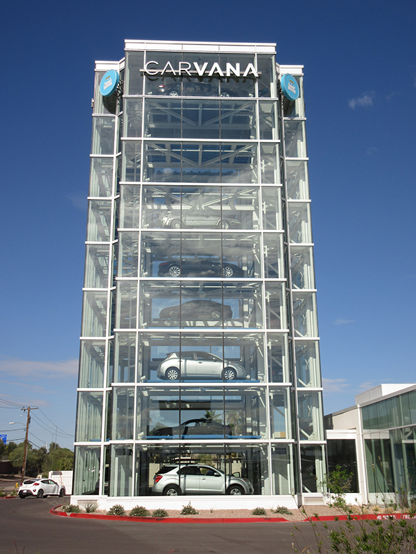 A tall, glass-walled Carvana car vending machine under a clear blue sky. Multiple cars are displayed on different levels inside the transparent structure, which resembles a modern vertical parking garage. The Carvana logo is prominently displayed at the top of the building, and the surrounding area includes pavement, landscaping, and part of an adjacent building.