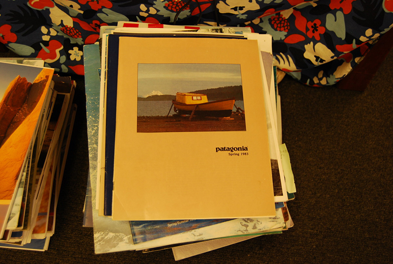 A stack of magazines or catalogs sits on the floor in front of a floral-patterned couch. The top catalog features a cover image of a yellow and red boat on a beach with mountains in the background. The cover is labeled "Patagonia Spring 1983." Additional colorful catalogs and publications are visible underneath and in a second stack to the left.