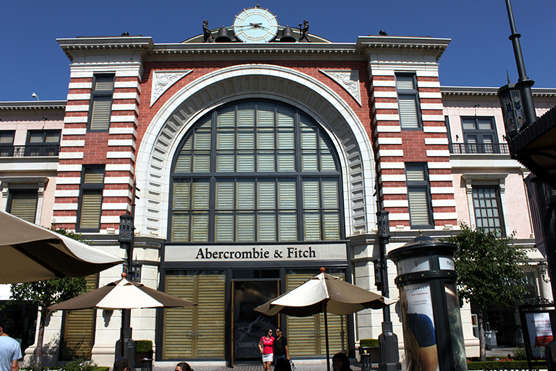 A large Abercrombie & Fitch store with a grand arched window in the center of its facade, featuring a mix of red brick and white accents. A clock is positioned at the top of the building. Several outdoor umbrellas and lampposts are visible in the foreground, along with a few people near the entrance. The scene is set on a sunny day with a clear blue sky
