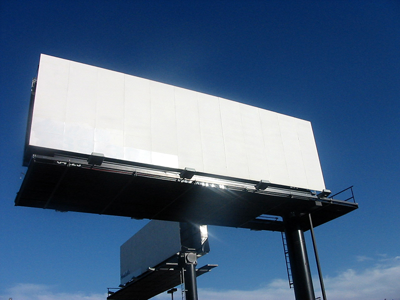 A low-angle shot of a large, rectangular, blank white billboard against a clear blue sky with a few wispy clouds. The billboard is mounted on black metal supports