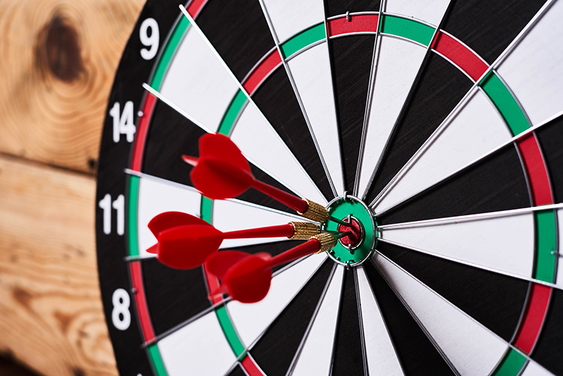 A close-up of a dartboard with three red darts embedded in the bullseye. The dartboard has black, white, green, and red segments, with numbers visible along the edge. The board is mounted on a wooden surface, and the image is taken at an angle, highlighting the precision of the darts hitting the center.