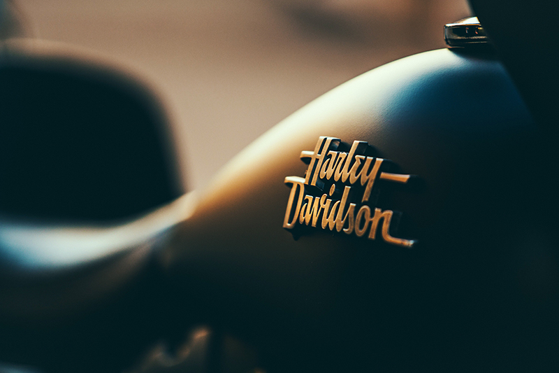 A close-up photograph of a Harley-Davidson motorcycle's fuel tank, featuring the brand's iconic logo in raised metallic lettering. The tank has a sleek, matte black finish with a smooth curve. The lighting in the image creates a warm, golden glow, emphasizing the contours of the bike and the details of the emblem. The background is blurred, drawing focus to the Harley-Davidson logo and the craftsmanship of the motorcycle.