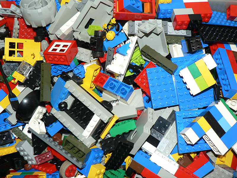 A large pile of assorted LEGO bricks and pieces in various colors, shapes, and sizes. The pile includes red, blue, yellow, green, black, white, and gray bricks, along with specialized pieces such as windows, wheels, and minifigure parts. Some pieces are connected, while others are scattered loosely, creating a chaotic and colorful arrangement.