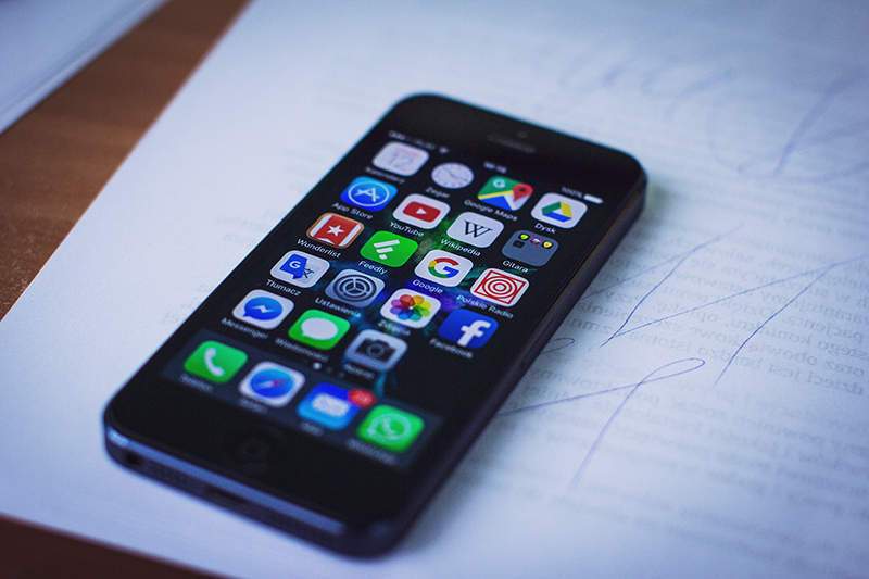 A smartphone with a black frame lies on a piece of paper with handwritten notes. The phone's screen is turned on, displaying a home screen filled with colorful app icons, including social media apps such as Facebook, Twitter, and WhatsApp, as well as utility apps like Google Maps, YouTube, and the App Store. The screen is reflective, and the background is slightly blurred, emphasizing the phone as the main subject.