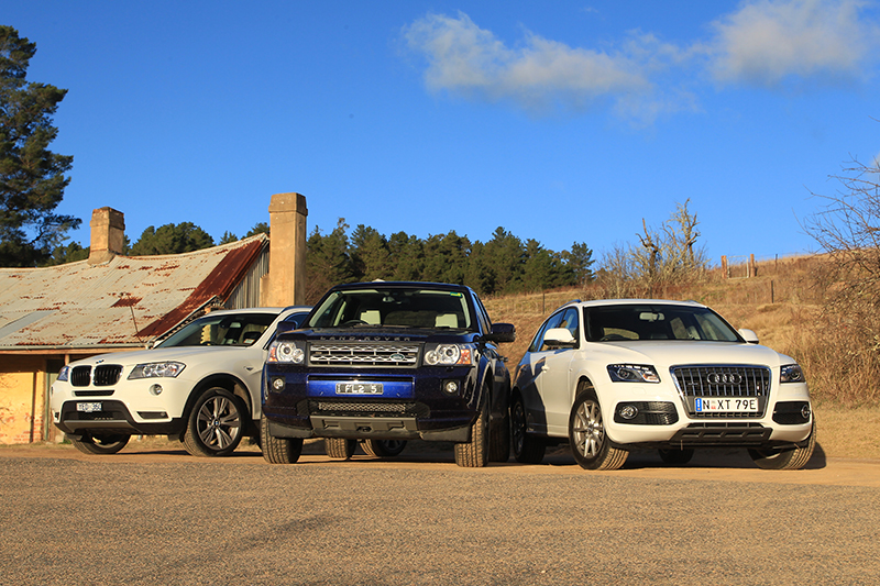Three SUVs are parked side by side on a paved surface in front of a rustic building with a rusted tin roof. The vehicles include a white BMW on the left, a black Land Rover in the center, and a white Audi on the right. In the background, there are trees, dry grass, and a clear blue sky with a few clouds