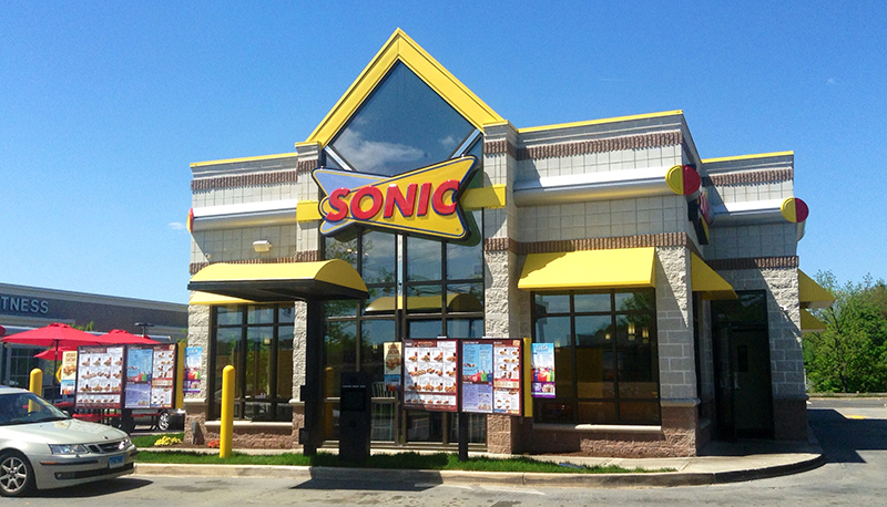 A Sonic Drive-In restaurant building under a clear blue sky. The structure features a modern design with large windows, yellow awnings, and a prominent red and yellow Sonic logo above the entrance. Multiple menu boards are visible outside, and a silver car is parked near the drive-thru ordering station