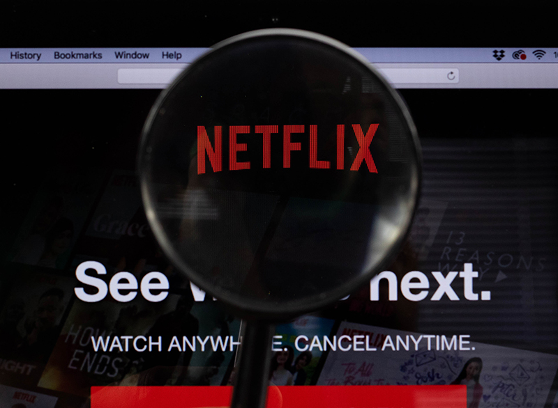 A close-up of a computer screen displaying the Netflix homepage, with a magnifying glass focusing on the red 'Netflix' logo. The background of the screen includes the text 'See what's next' and 'Watch anywhere. Cancel anytime.' partially visible. The magnifying glass enlarges the 'Netflix' logo, making it stand out against the dark background.