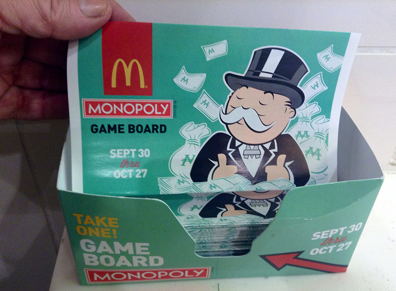 A close-up of a McDonald's Monopoly game board display box. The box contains folded game boards and features the Monopoly mascot, Rich Uncle Pennybags, surrounded by dollar bills and money bags. Text on the game board and box reads “MONOPOLY GAME BOARD” with promotional dates “Sept 30 thru Oct 27.” A hand is seen picking up one of the game boards.