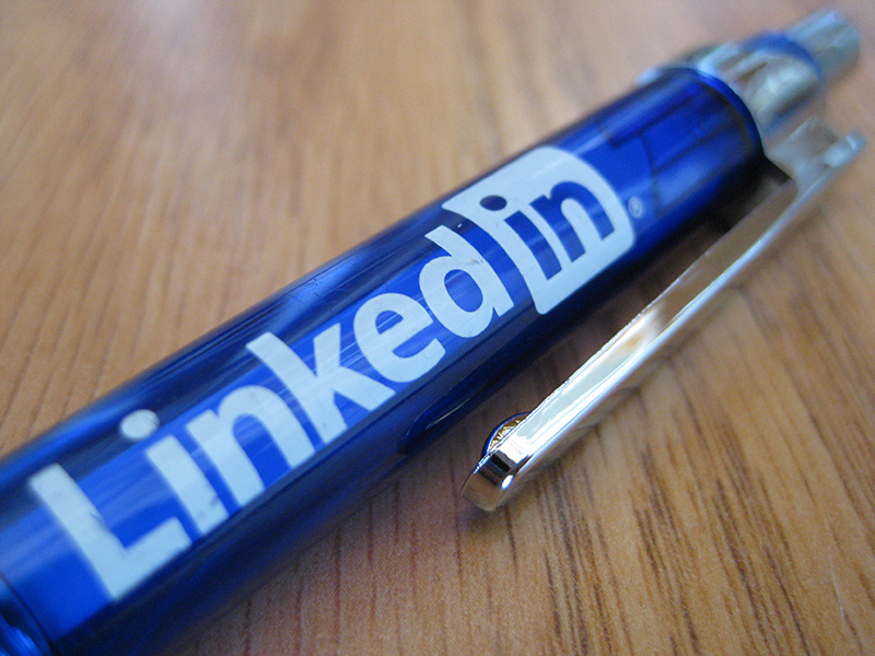 A close-up of a blue pen with the LinkedIn logo printed in white letters on the barrel. The pen rests on a wooden surface, and the metal clip is clearly visible along the side.