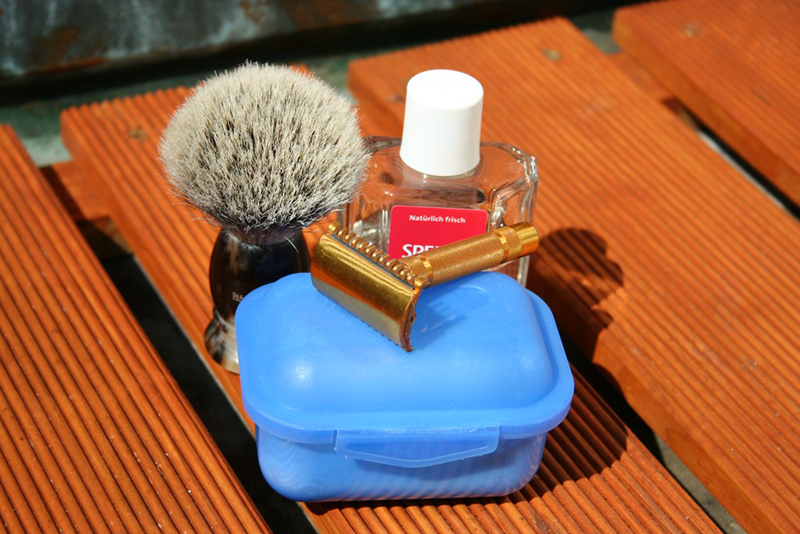 A traditional shaving kit displayed on a wooden surface. The kit includes a shaving brush with a dark handle and light bristles, a bottle of aftershave with a white cap labeled in German, a golden safety razor, and a blue plastic soap container with a lid. Sunlight casts shadows across the items
