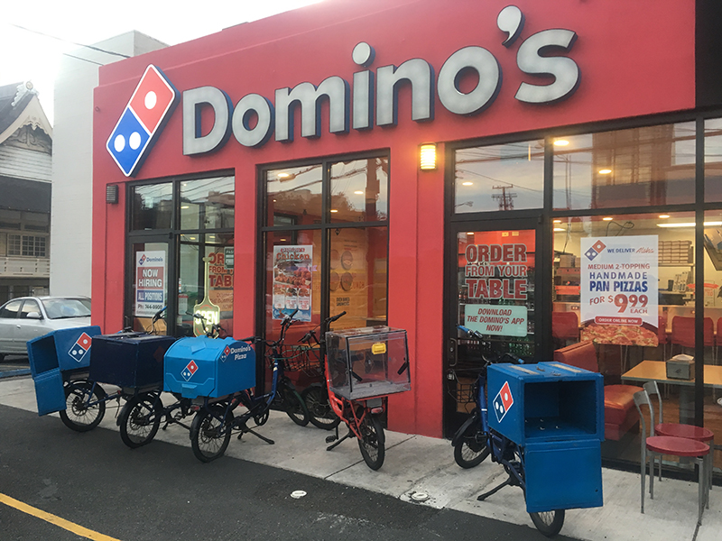 The exterior of a Domino’s Pizza restaurant with a bold red facade and large white Domino’s logo above the entrance. Several delivery bicycles with blue and red insulated cargo boxes are parked outside on the sidewalk. Signs in the windows advertise job openings, mobile ordering, and a pizza promotion for $9.99. The interior shows red seating and lit signage