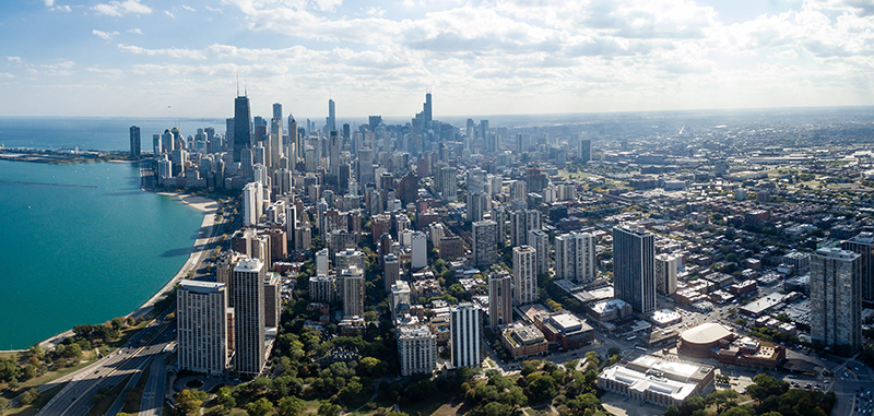 Aerial view of a large city skyline bordered by a lake. The city is densely packed with tall skyscrapers and mid-rise buildings, gradually transitioning into residential areas. The lake, seen on the left, has a curved shoreline with a highway running alongside it. The scene is brightly lit under a partly cloudy sky.