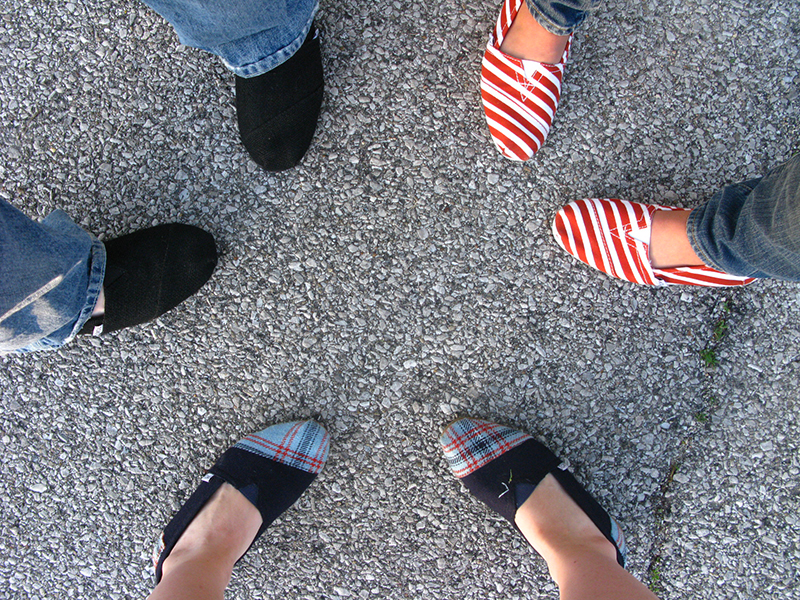A top-down view of four people standing in a circle on a gravel surface, with only their feet visible. Each person is wearing different casual slip-on shoes: one pair is black, another red and white striped, and two pairs are plaid with blue and red patterns
