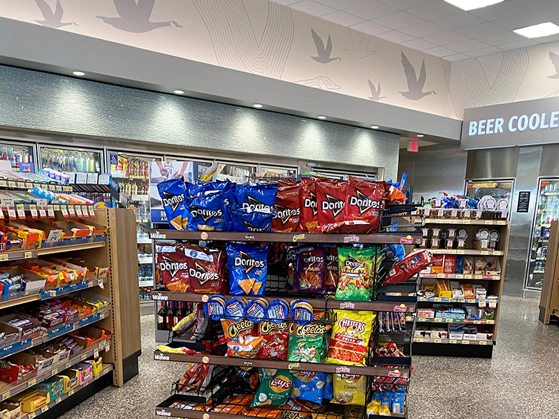 An interior view of a convenience store showing neatly arranged shelves stocked with various snack products. The central display features large bags of Doritos, Cheetos, Lay’s, and other chips. Surrounding shelves display candy, packaged snacks, and grocery items. In the background, there are refrigerated beverage coolers and a sign labeled “Beer Cooler” above a doorway. The ceiling has recessed lighting and a decorative mural of flying birds