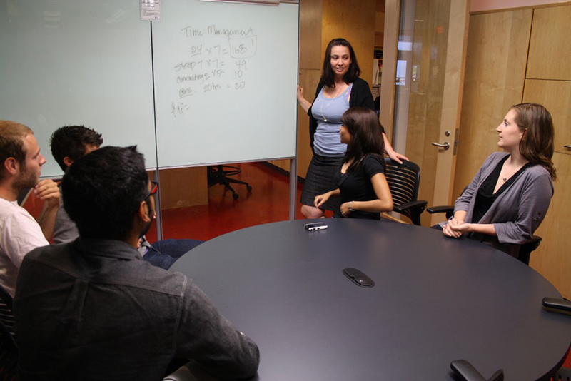 A group of five people in a modern conference room engaged in a meeting. A woman is standing near a whiteboard, explaining a topic labeled 'Time Management,' with various calculations written on the board. Four other individuals, two women and two men, are seated around a large black conference table, attentively listening. The room has glass walls, wooden cabinets, and office chairs, creating a professional workspace atmosphere.