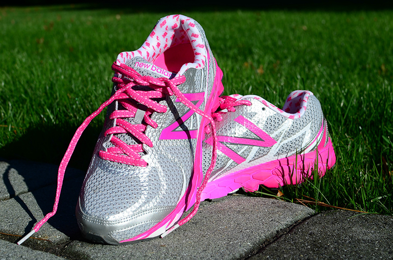 A pair of silver athletic running shoes with bright pink accents, including laces, soles, and design details. The shoes, branded with the New Balance logo, are placed on a stone path with green grass in the background, brightly lit by sunlight.