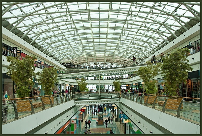 A spacious, multi-level indoor shopping mall with a large curved glass ceiling allowing natural light to fill the area. The mall features modern architecture with clean lines and metal railings. Several green plants and trees are placed along the walkways on the upper floors. Shoppers can be seen walking and browsing stores on each level.