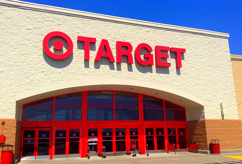 The exterior of a Target retail store under a clear blue sky. The building features a large red Target logo and bold red letters spelling “TARGET” above a wide entrance with red-framed glass doors and windows. Shopping carts and red bollards are visible near the entrance.